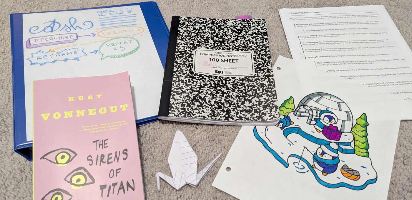 A composition notebook, blue binder, paper crane, list of cognitive distortions, coloring page of penguins in snow, and a paperback copy of "Sirens of Titan" by Kurt Vonnegut all on carpet