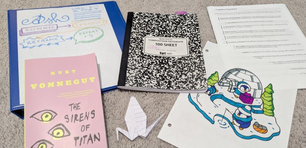 A composition notebook, blue binder, paper crane, list of cognitive distortions, coloring page of penguins in snow, and a paperback copy of "Sirens of Titan" by Kurt Vonnegut all on carpet